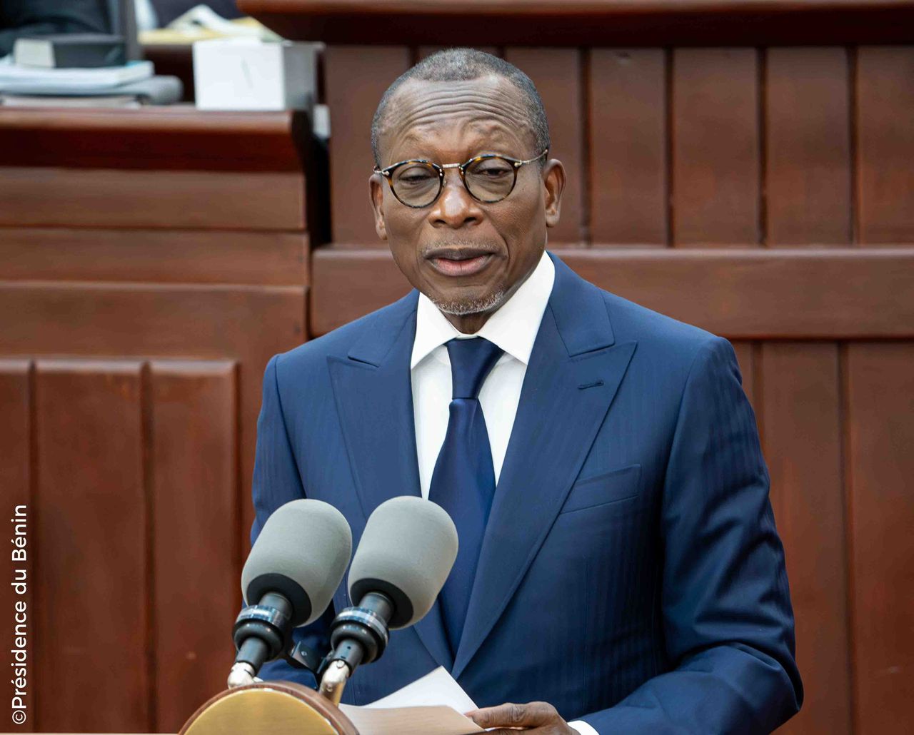Homme en costume bleu et lunettes parlant devant deux micros dans un cadre officiel en bois brun