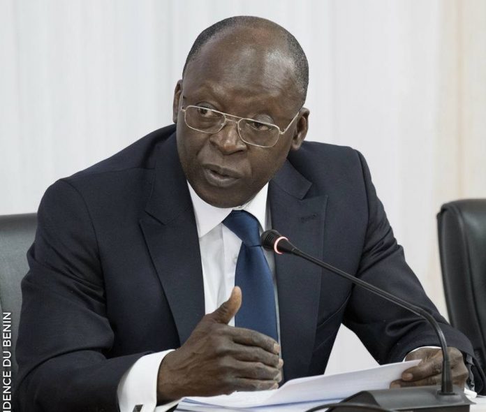 Homme en costume sombre et cravate bleue parle face à un microphone en tenant des documents au cours d'une réunion