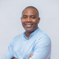 Un homme souriant en chemise bleue claire, bras croisés, posant devant un fond uni gris clair