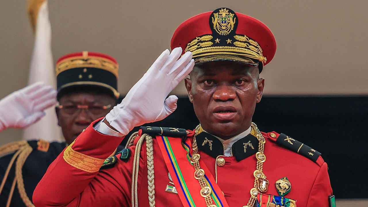 Un homme en uniforme militaire rouge avec plusieurs décorations salue de la main droite levée en gant blanc