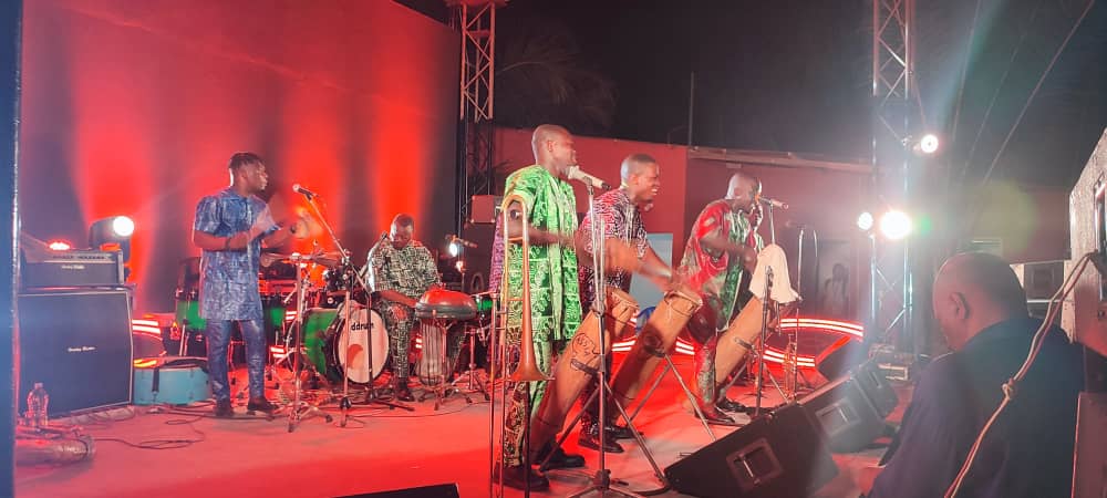 Quatre musiciens jouent des percussions et deux autres chantent sur une scène éclairée en rouge devant un public