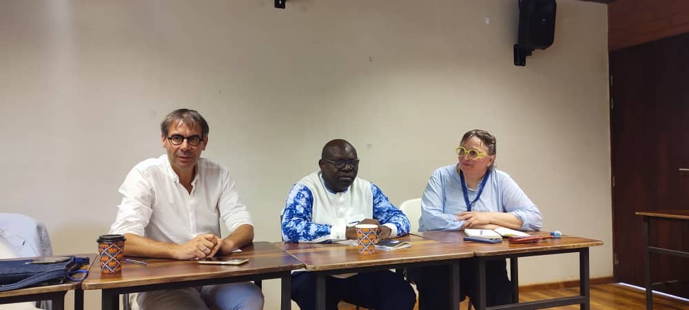 Trois personnes assises à une table face au public dans une salle de conférence avec mic et gobelets colorés.