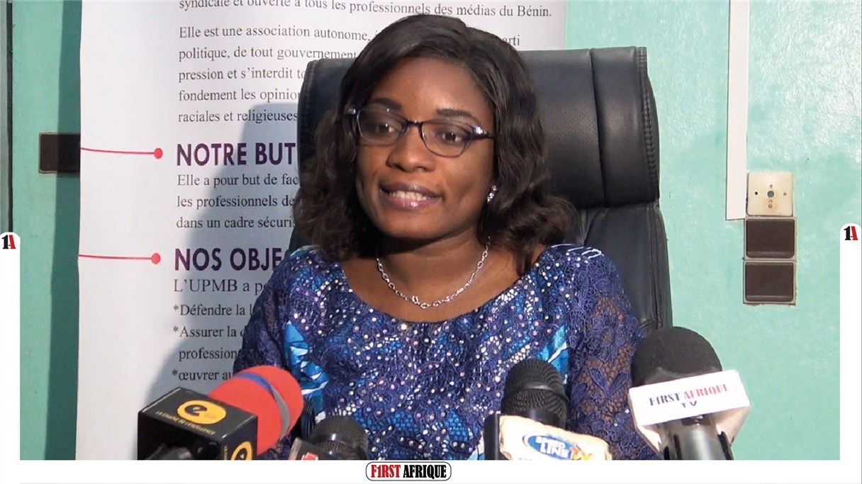 Une femme souriante en robe bleue parle devant plusieurs micros dans un bureau avec un panneau d’informations en arrière-plan