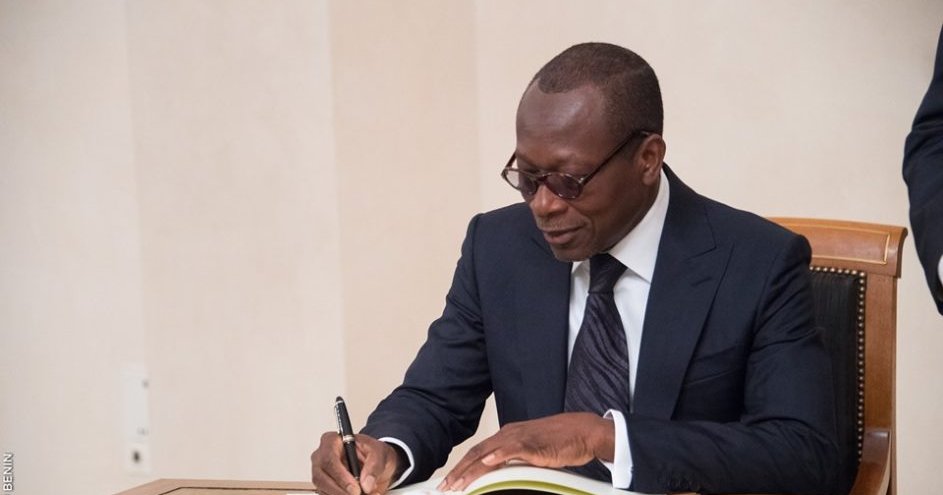Un homme en costume noir et lunettes signe un document posé sur une table devant un mur blanc