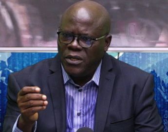 Homme portant des lunettes noires et costume sombre parlant en gesticulant avec la main droite devant un micro