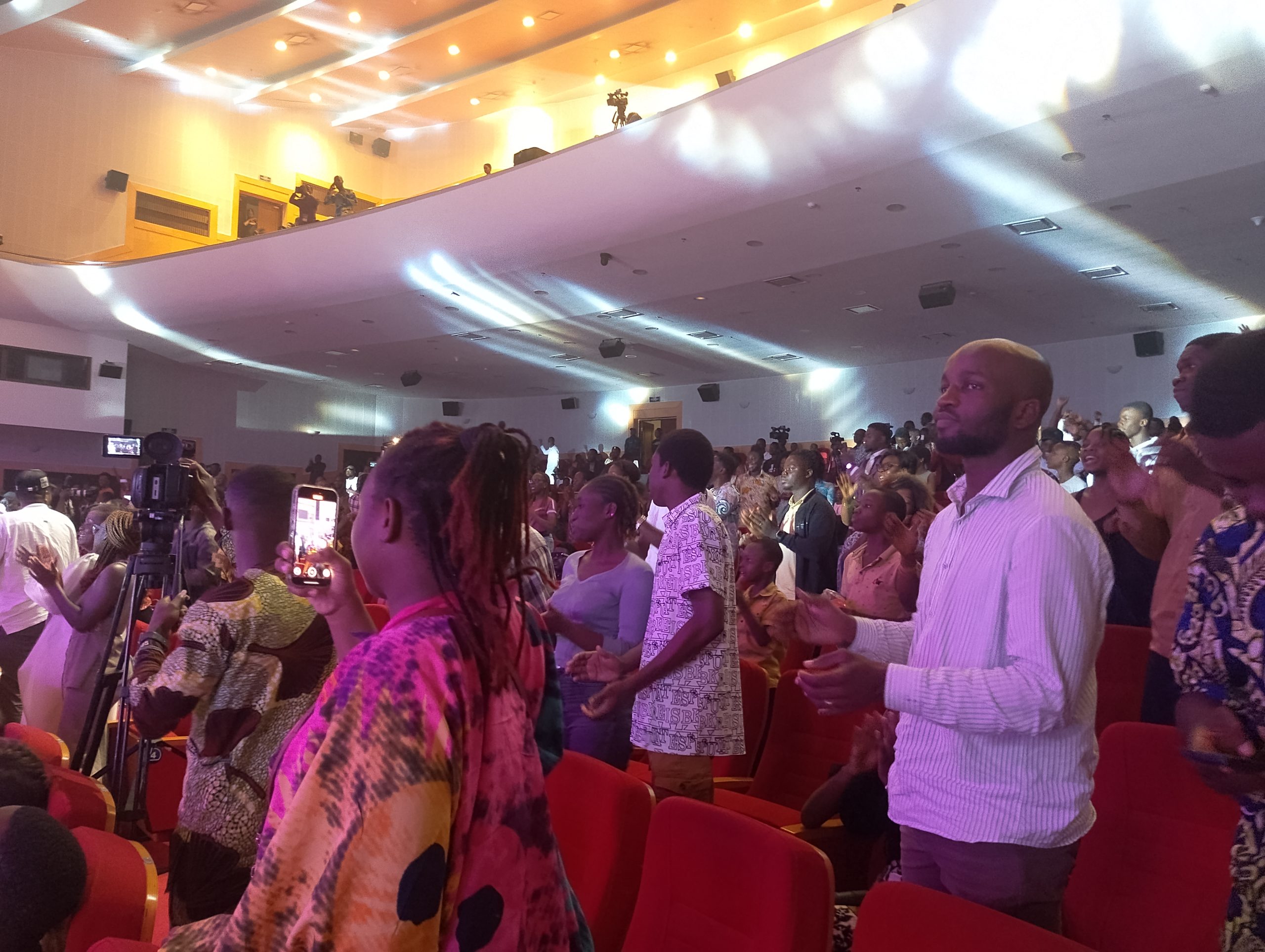 Personnes debout dans une salle de spectacle éclairée, certaines filment ou applaudissent, ambiance de rassemblement culturel