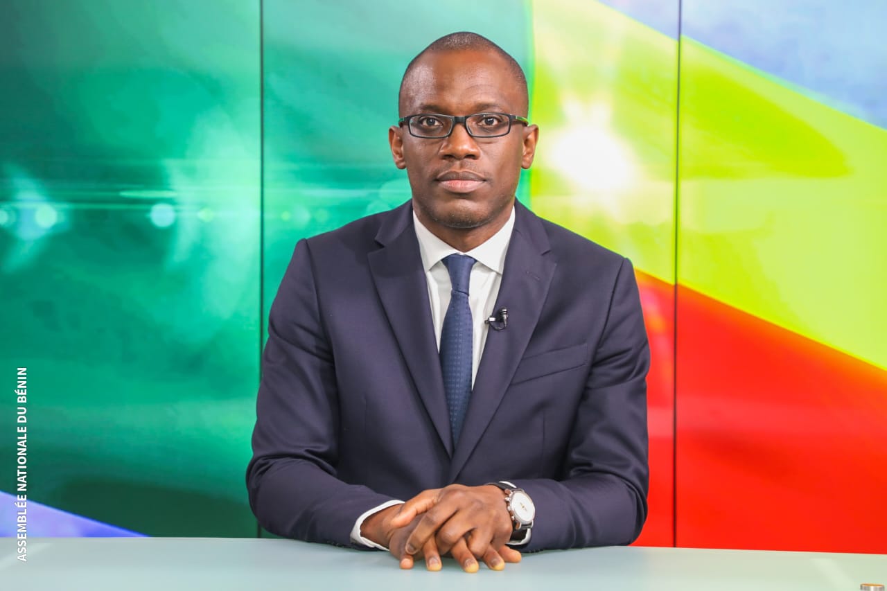 Un homme en costume sombre et cravate, portant des lunettes, est assis derrière un bureau avec un fond coloré vert, jaune et rouge.