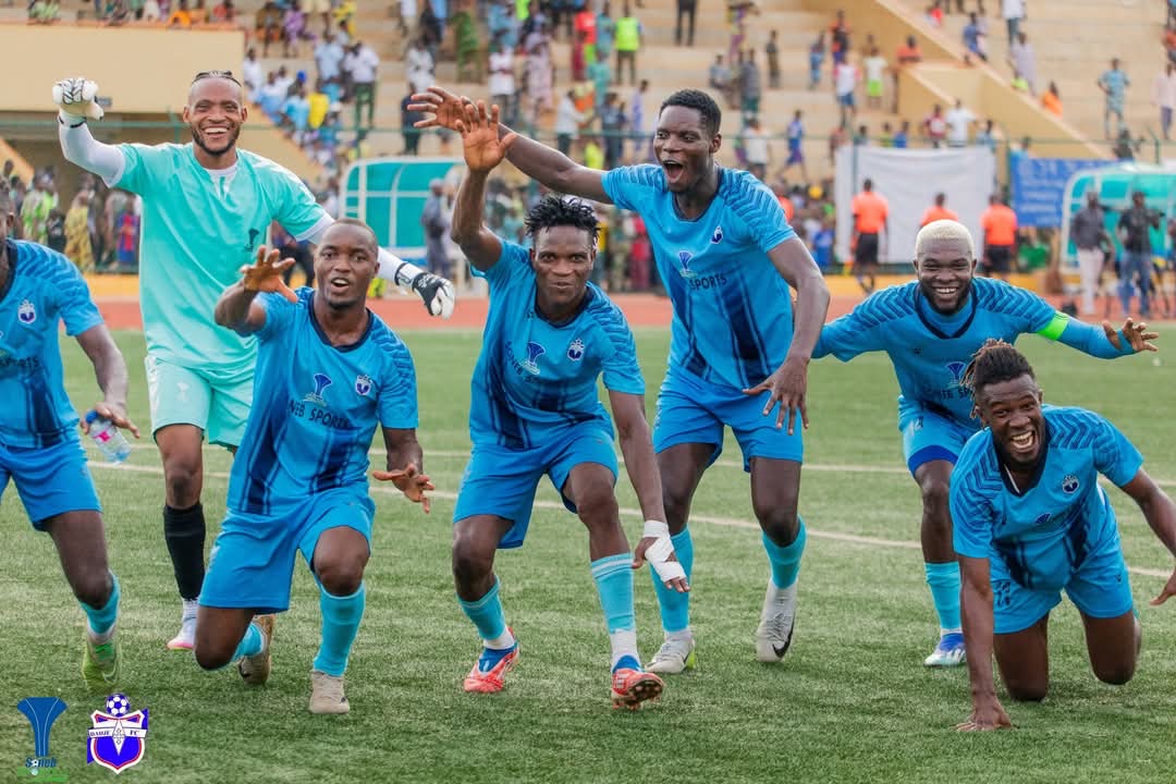 six joueurs de football en tenue bleue posent et sourient sur un terrain devant un public dans les tribunes