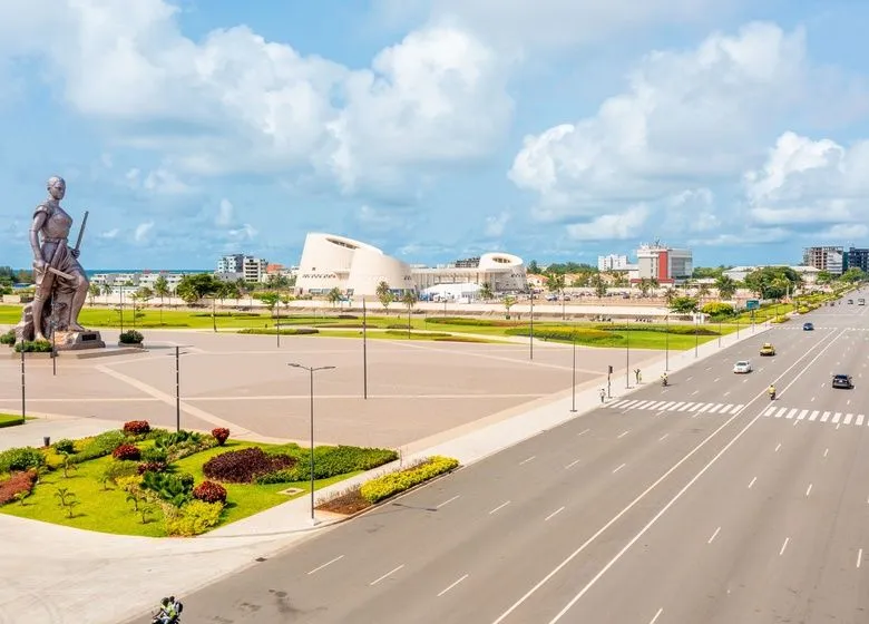 Grand boulevard urbain désert avec un grand espace vert, une statue monumentale et un bâtiment moderne en fond sous un ciel bleu nuageux