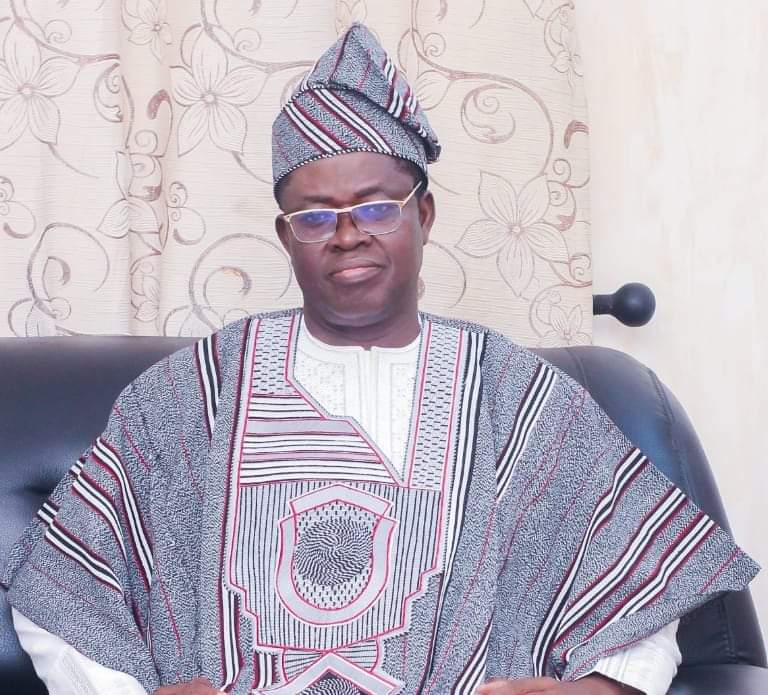 Homme assis sur un canapé en tenue traditionnelle africaine rayée avec chapeau assorti et lunettes devant un rideau fleuri beige