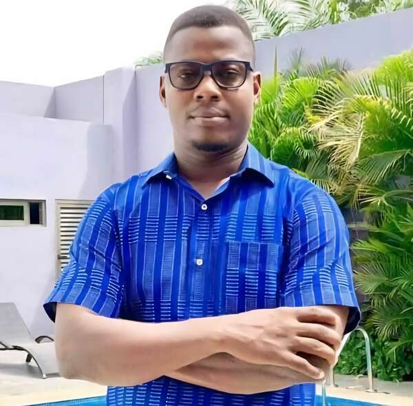 Homme portant une chemise bleue à motifs, lunettes, bras croisés devant une piscine et un mur clair avec des plantes vertes