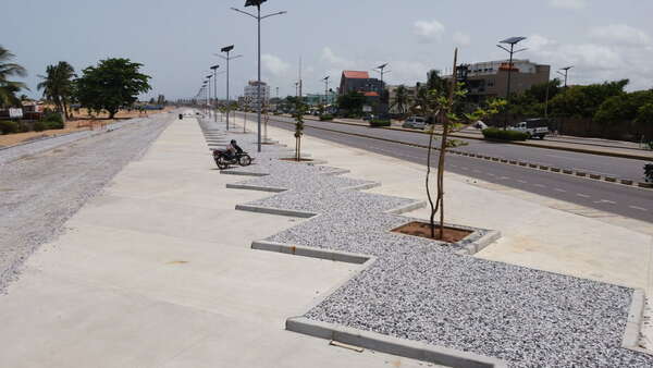 Trottoir large avec emplacements délimités par gravier et jeunes arbres bordant une route sous un ciel partiellement nuageux