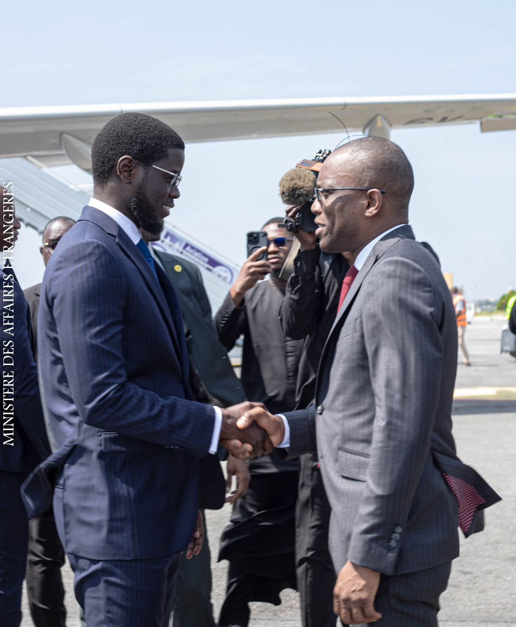 Deux hommes en costume s’accordent la main devant un avion sur un tarmac avec plusieurs personnes en arrière-plan