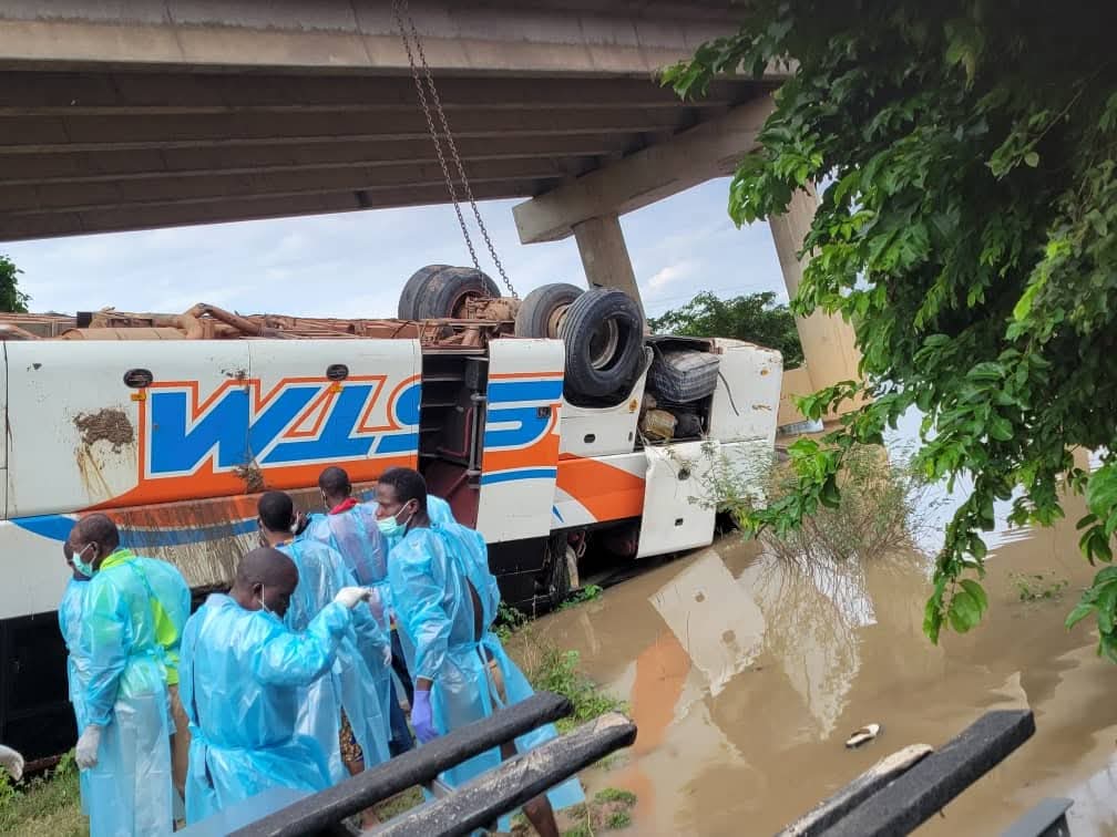 Un bus renversé sur le côté dans l'eau sous un pont avec plusieurs personnes en tenues de protection bleues autour