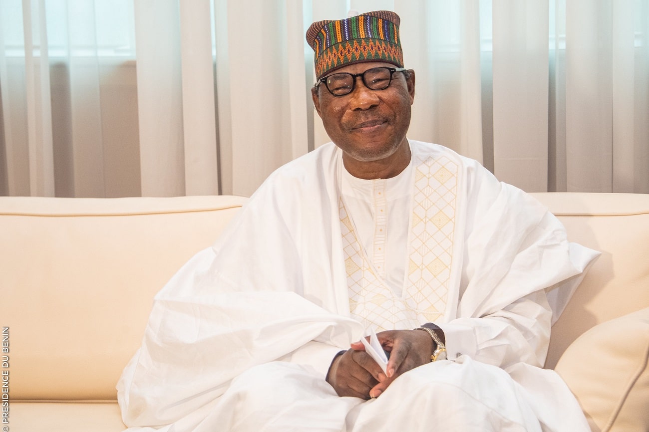 Homme souriant en tenue traditionnelle blanche et chapeau coloré assis sur un canapé beige devant un rideau blanc transparent