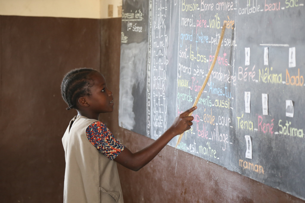 Une élève tient un bâton et montre des mots écrits à la craie colorée sur un tableau noir dans une salle de classe.