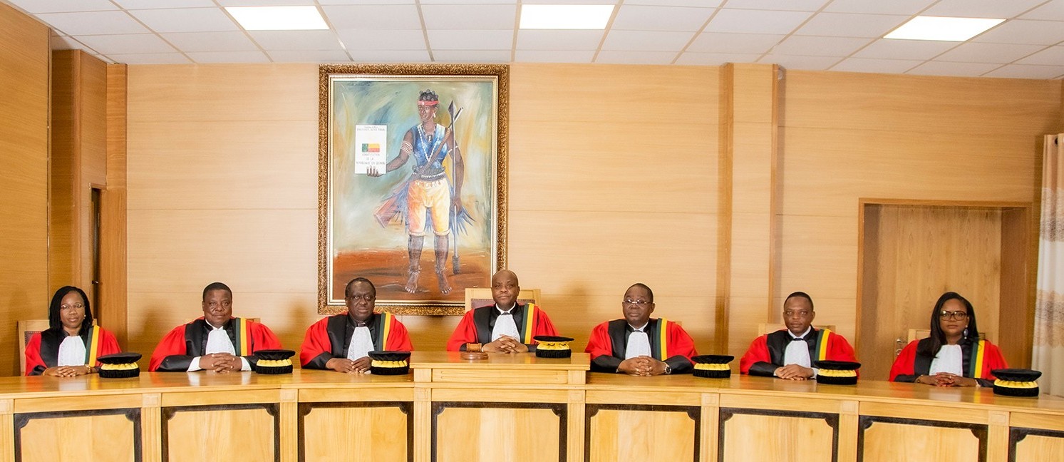 Sept membres en robe rouge et noire assis derrière un bureau en bois dans une salle ornée d'un grand portrait encadré
