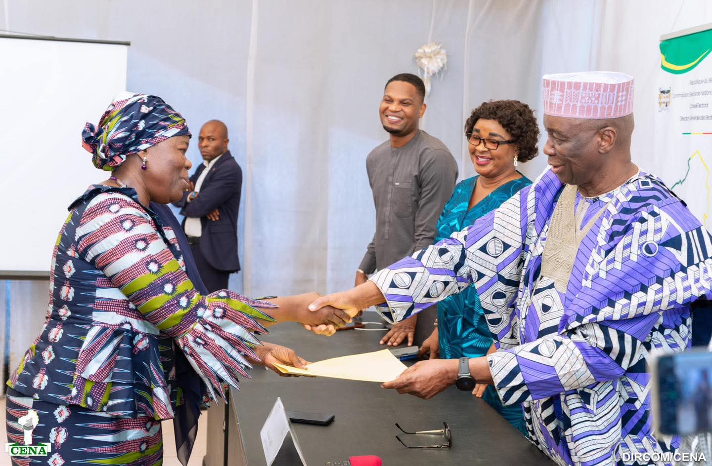 Un homme remet un document à une femme habillée en tenue traditionnelle colorée lors d’une cérémonie officielle avec d’autres personnes souriantes à table