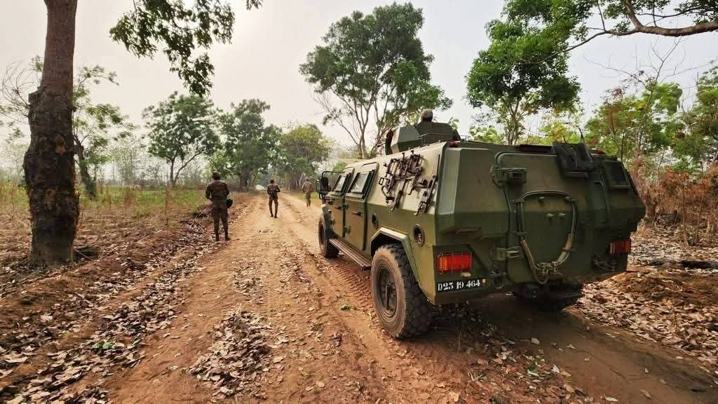 Véhicule blindé militaire sur une piste en terre entourée d’arbres avec trois soldats marchant à proximité