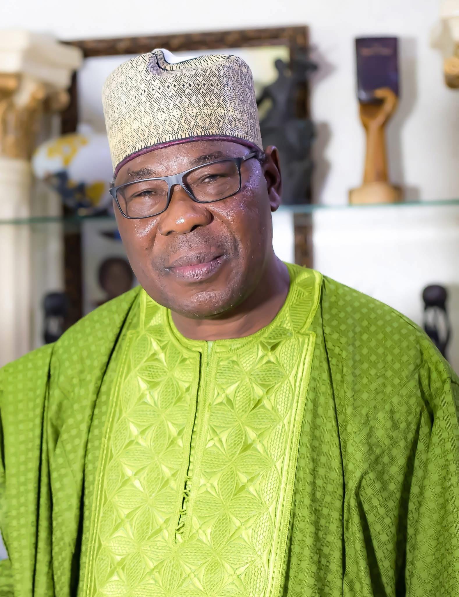 Homme portant un boubou vert et un chapeau traditionnel beige avec des lunettes debout devant une étagère avec objets décoratifs