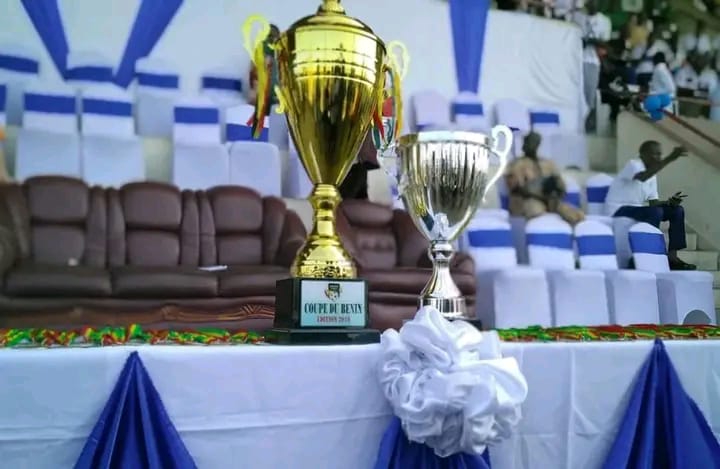 Deux trophées posés sur une table décorée avec des tissus bleu, blanc et vert dans une tribune de stade vide partiellement occupée