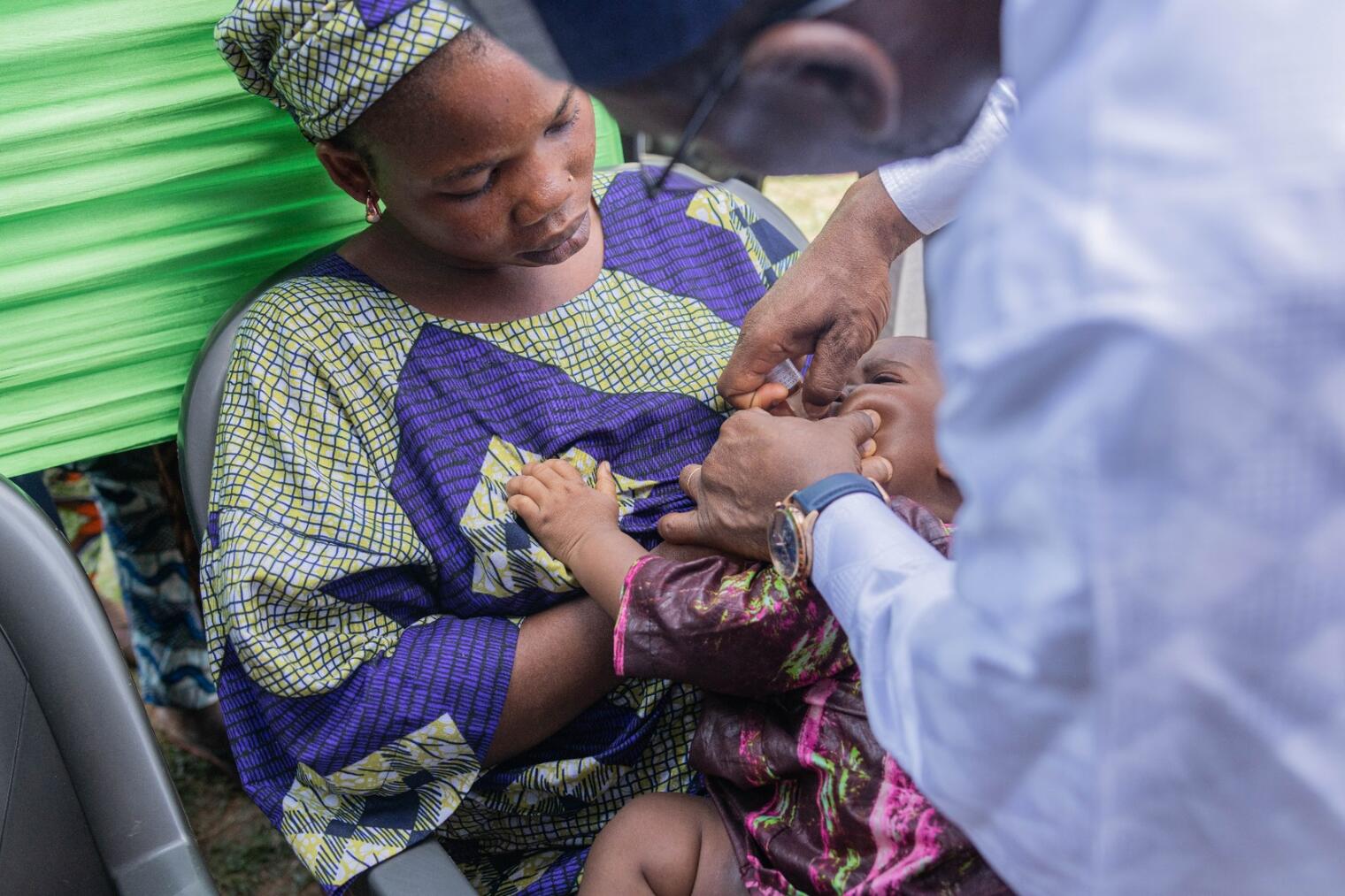 Une femme en tenue traditionnelle tient un bébé tandis qu'un soignant lui donne un vaccin oral contre la poliomyélite