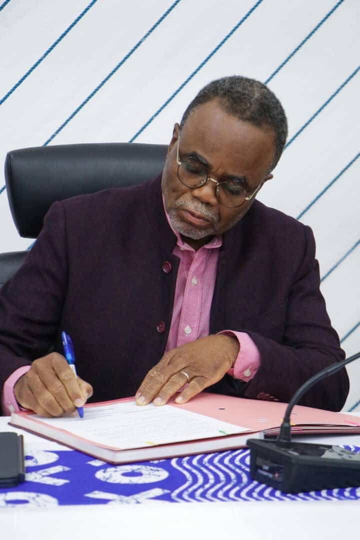Un homme en veste sombre signe un document posé sur une table recouverte d’un tissu à motifs bleus et blancs