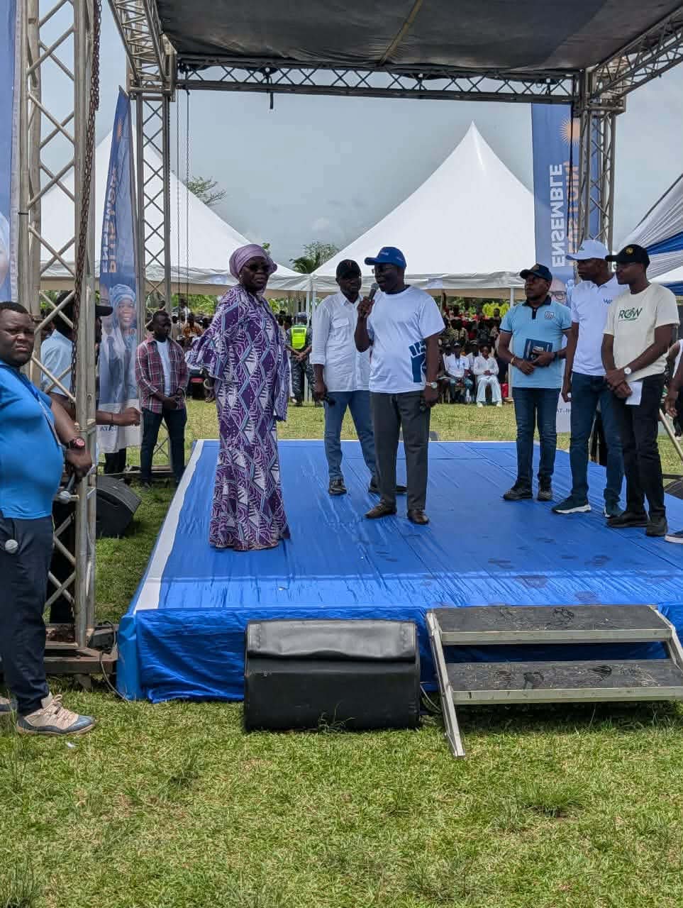 Personnes debout sur une estrade bleue sous une structure métallique avec un micro, public et tentes blanches en arrière-plan