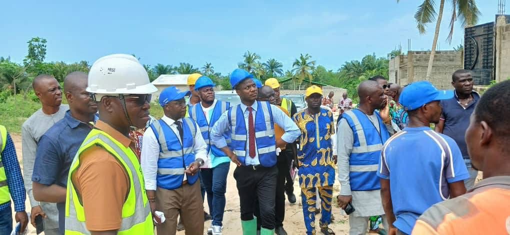 Un groupe d'hommes en tenue de chantier et bavette bleue discute sur un terrain sablonneux sous un ciel bleu.