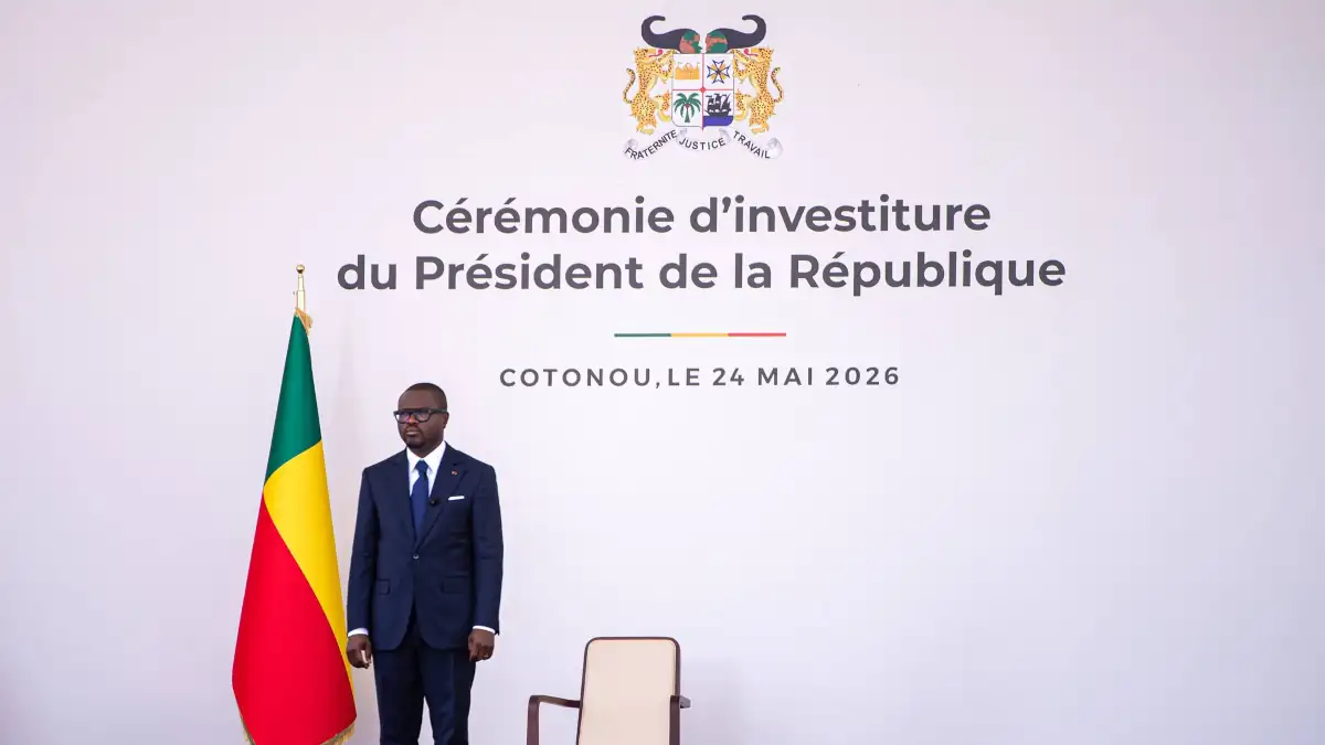 Un homme en costume sombre et cravate bleue se tient debout à côté d'un drapeau aux couleurs vert, jaune et rouge devant un mur blanc avec un texte officiel et