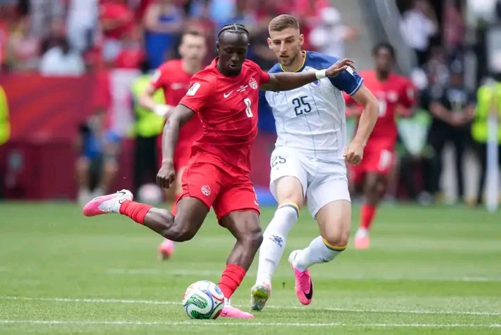 Deux joueurs de football en pleine action, l'un en tenue rouge frappant le ballon, l'autre en blanc le défiant sur le terrain vert