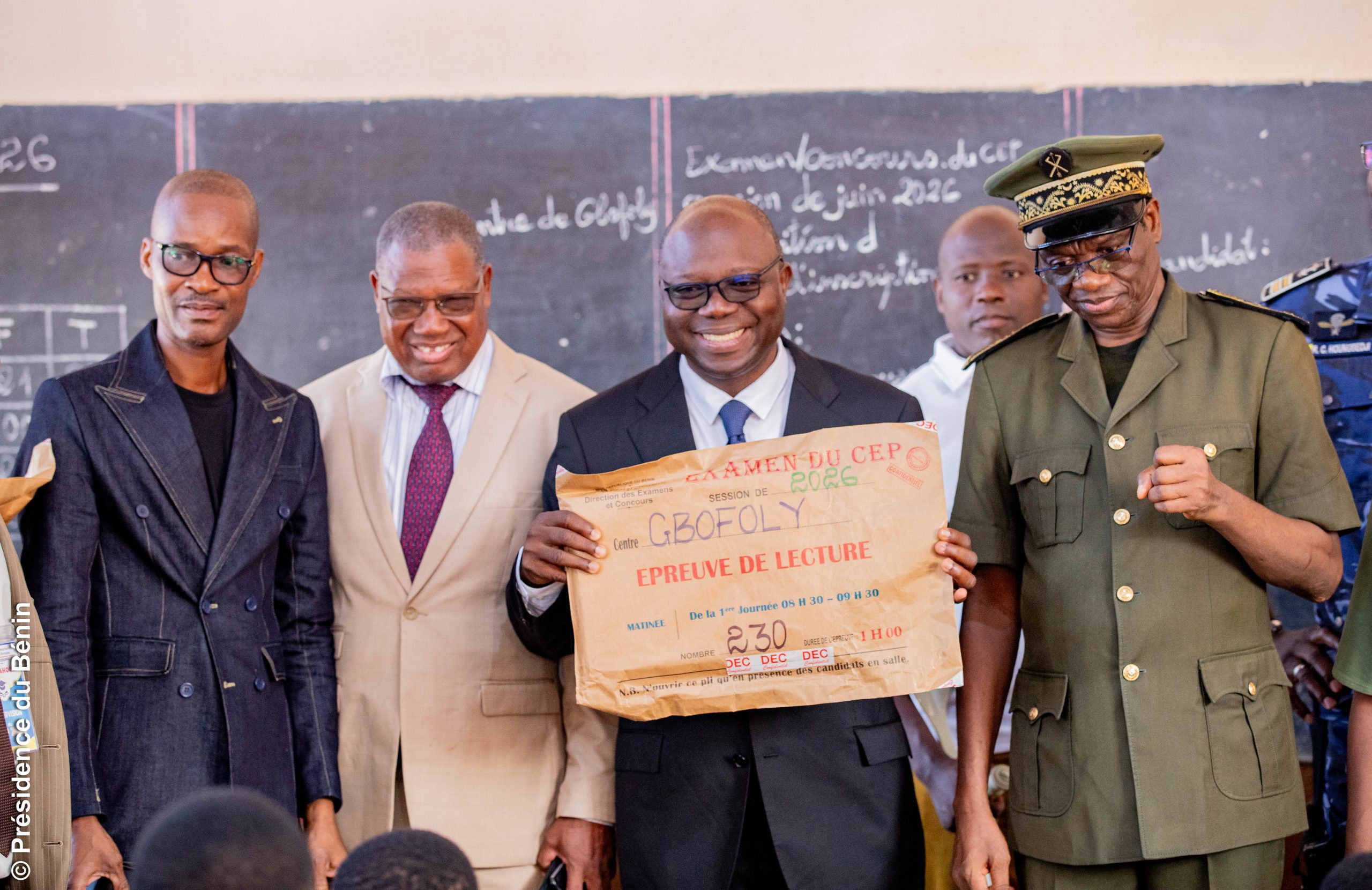 Quatre hommes souriants dont un en uniforme posent devant un tableau noir avec un grand papier indiquant une épreuve de lecture