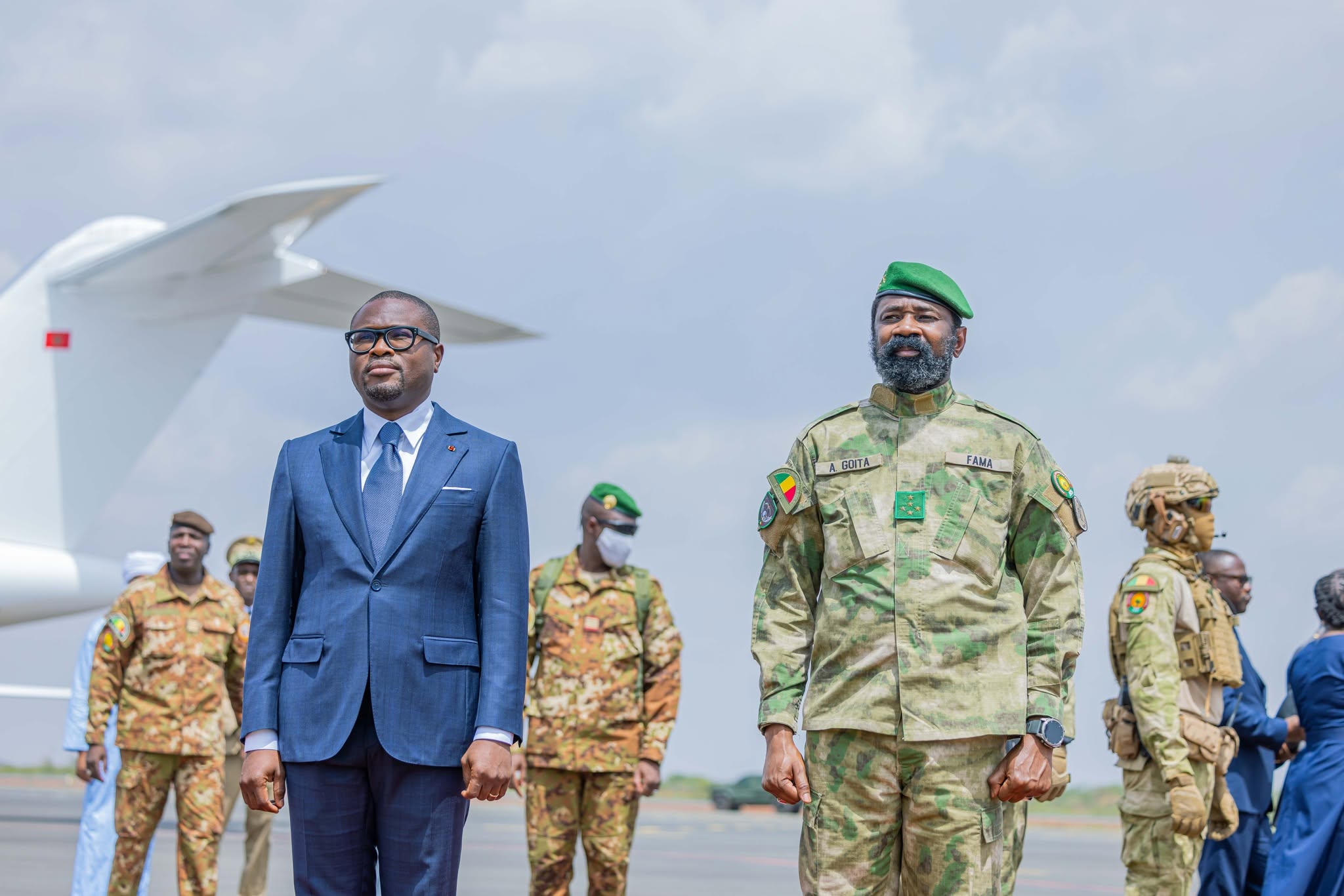 Deux hommes debout côte à côte, l’un en costume bleu et l’autre en tenue militaire verte, avec plusieurs soldats en arrière-plan sur un tarmac d’aéroport
