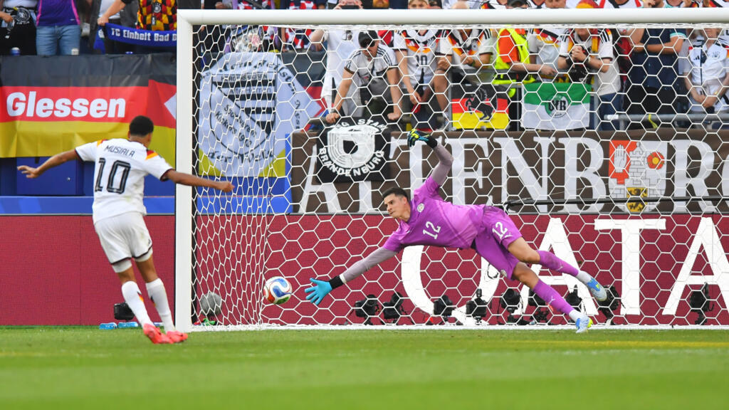 Un joueur en maillot blanc tire un penalty face au gardien en tenue rose plongeant pour arrêter le ballon