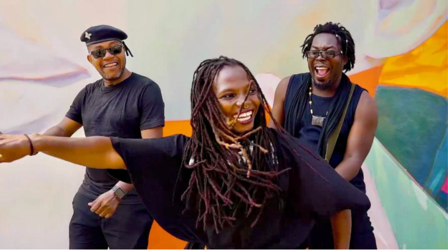 Trois personnes souriantes, deux hommes et une femme aux dreadlocks, danse devant un mur coloré abstrait