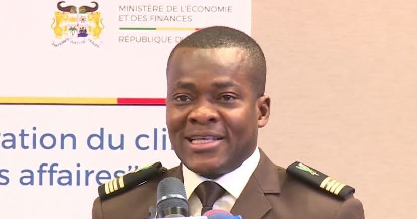 Un homme en uniforme militaire parle devant un microphone avec un panneau officiel en arrière-plan.