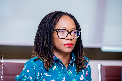 Une femme aux cheveux tressés et lunettes noires porte une chemise bleue à motifs floraux assise devant un mur clair