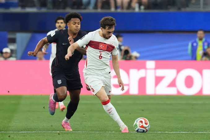 Deux joueurs de football en plein match, l'un en maillot blanc avec un drapeau rouge sur la poitrine contrôle le ballon devant un adversaire en maillot noir