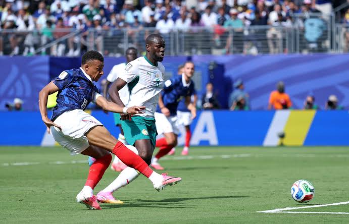 Un joueur en maillot bleu et short blanc tire un ballon alors qu’un adversaire en maillot blanc tente de l’en empêcher sur un terrain de football devant des spe