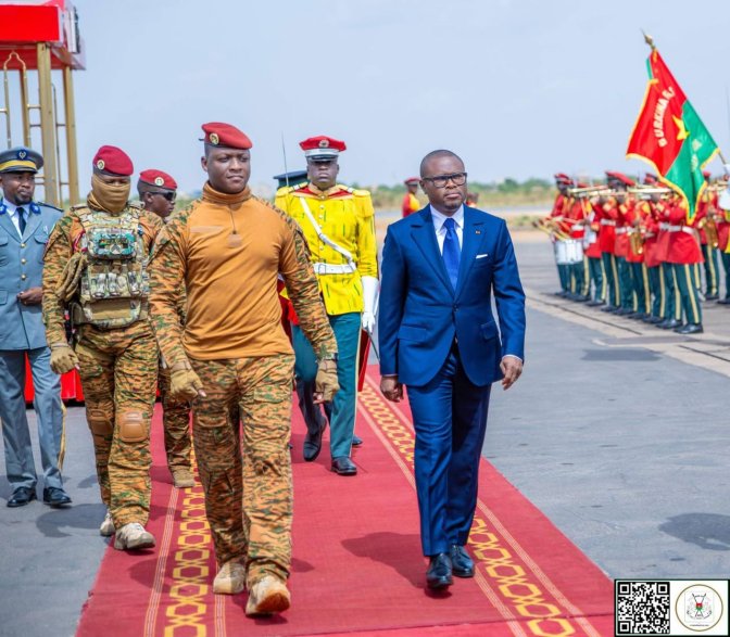 Un homme en costume bleu marche sur un tapis rouge suivi par des militaires en tenue camouflée et casques rouges devant une garde d'honneur.