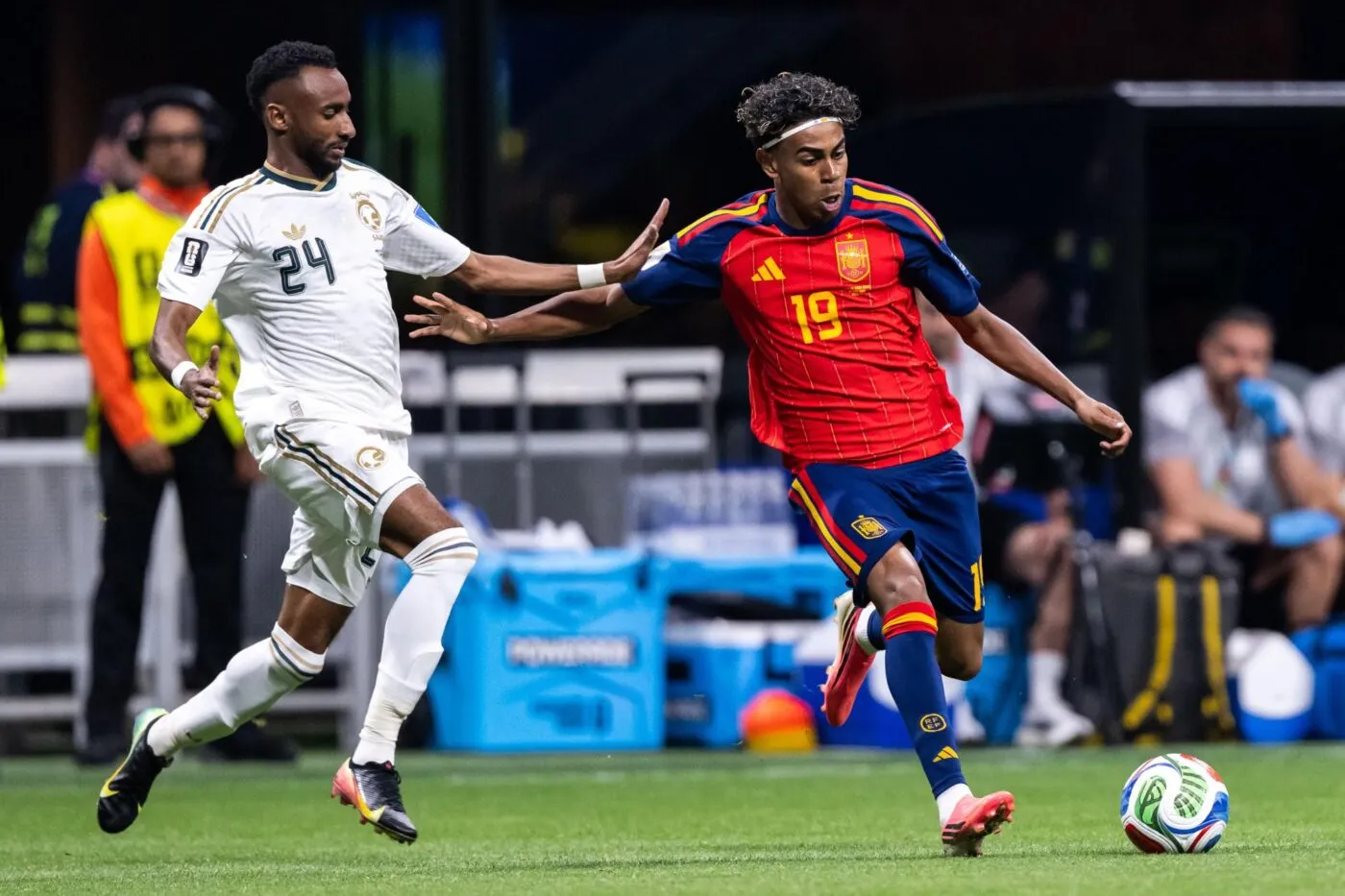 Deux joueurs de football en action, l’un en maillot rouge et bleu contrôle le ballon face à un adversaire en blanc