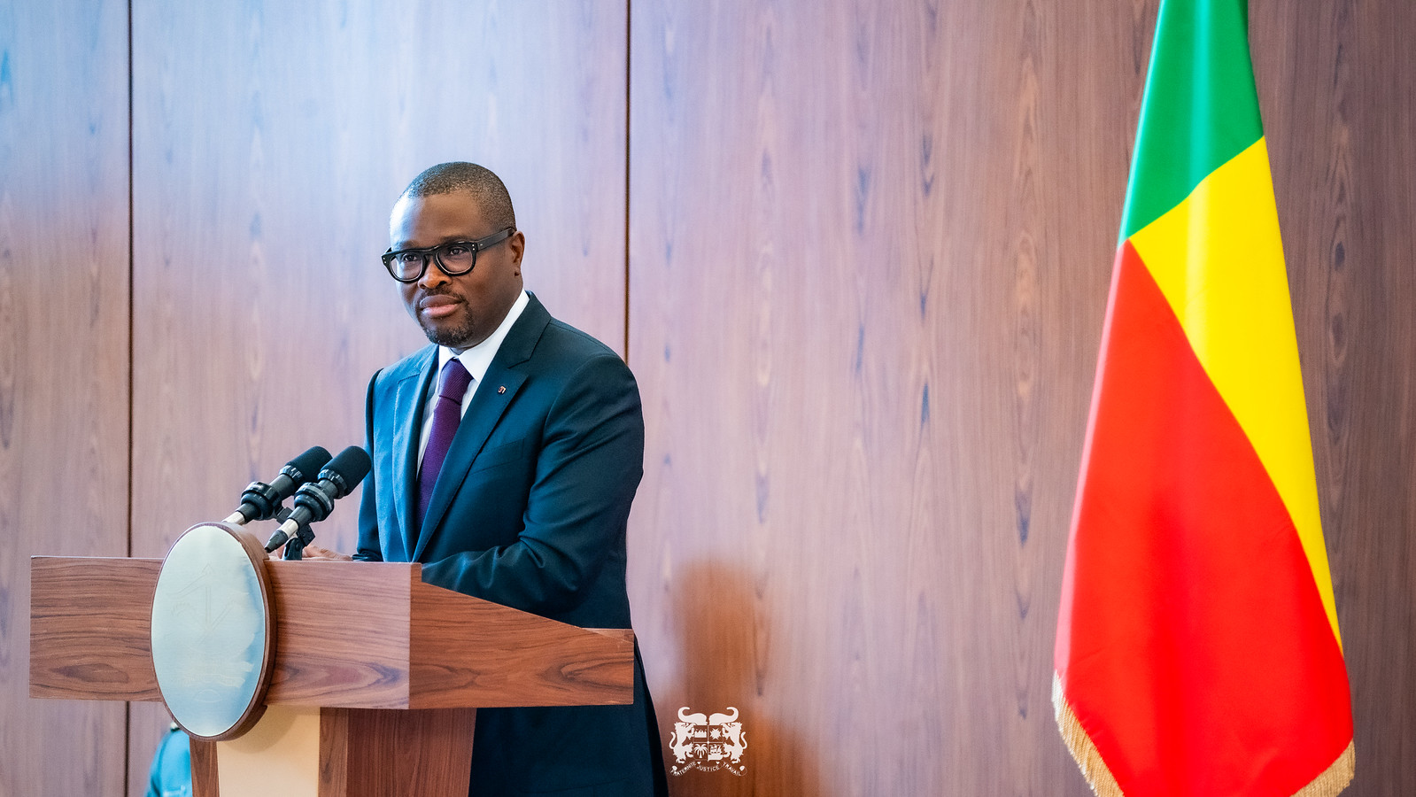 Homme en costume sombre et lunettes parlant derrière un pupitre en bois à côté d un drapeau tricolore vert jaune rouge
