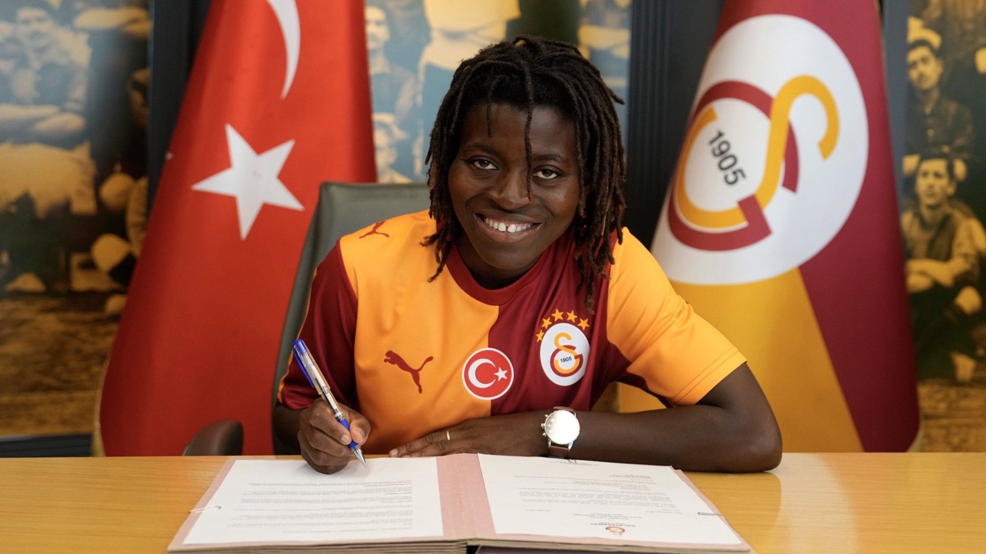 Jeune femme souriante en maillot rouge et jaune signe un contrat face à des drapeaux turc et de club sportif
