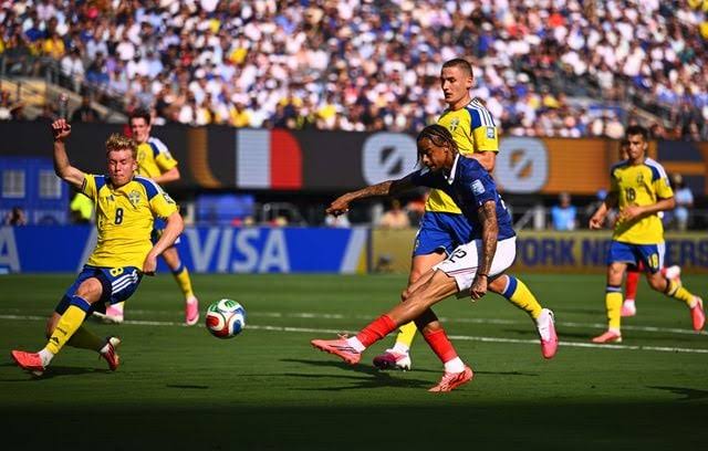 Un joueur en maillot bleu et blanc tire le ballon face à plusieurs adversaires en maillot jaune sur un terrain de football devant un stade rempli